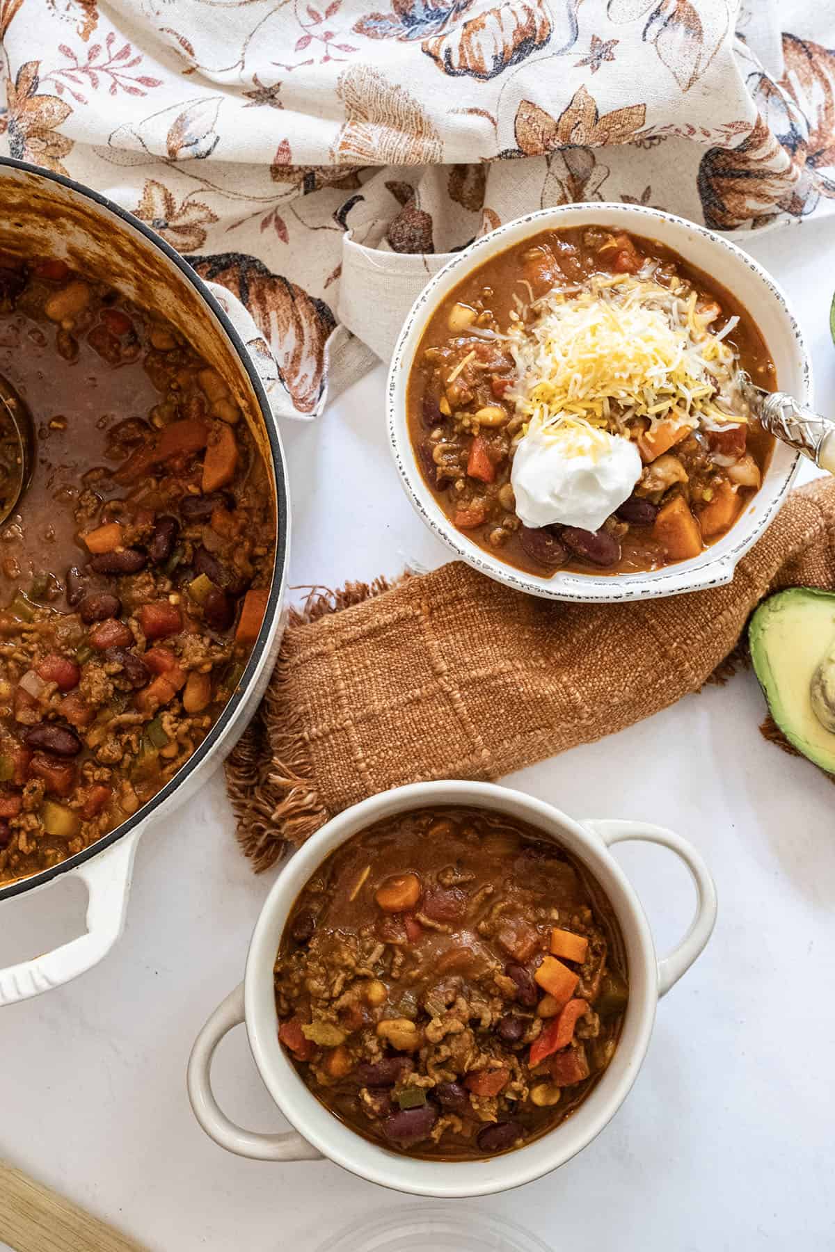 2 bowls and a dutch oven full of pumpkin chili with one bowl having sour cream and cheese added.