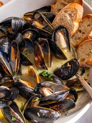 Steamed mussels in a serving bowl filled with white wine sauce and crostini on the side.