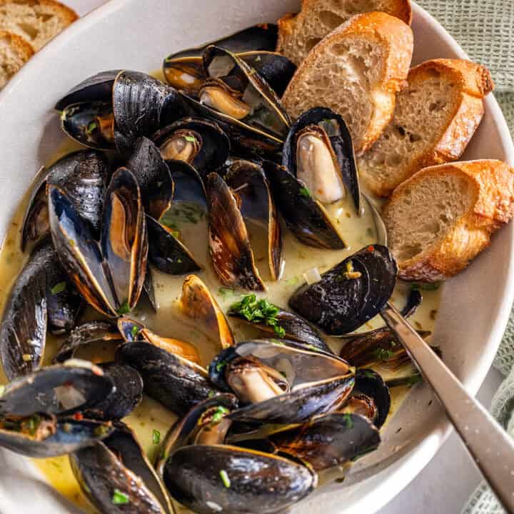 Steamed mussels in a serving bowl filled with white wine sauce and crostini on the side.
