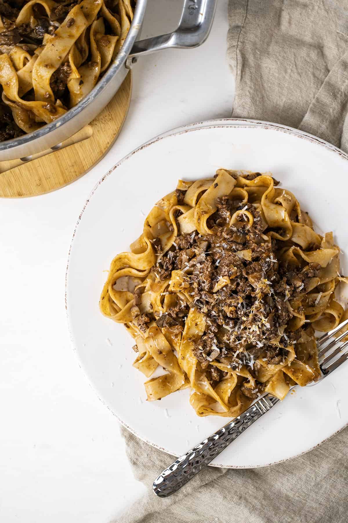 Lamb ragu over pappardelle on a small plate with pan with pasta next to it.