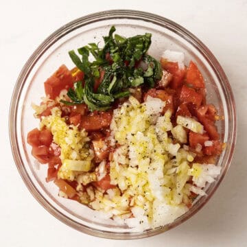 Tomatoes with onion, garlic, basil, olive oil, salt and pepper, in a bowl.