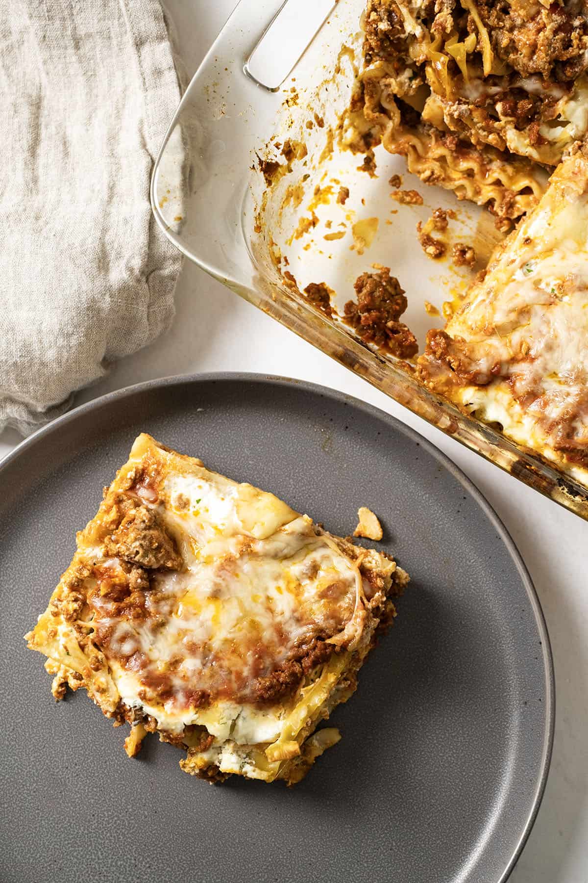 Square of lasagna on a plate with a baking dish filled with lasagna.