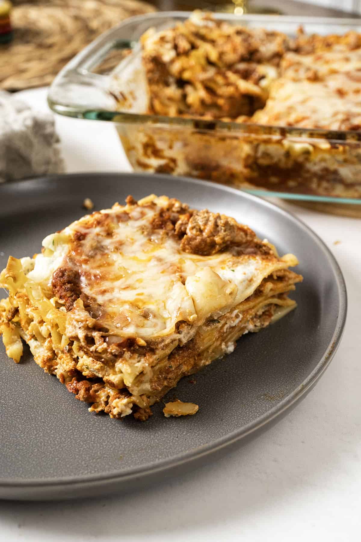 Square of lasagna on a plate with a baking dish filled with lasagna.