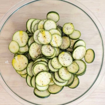 Zucchini slices in a bowl drizzled with olive oil and seasoned with salt and pepper.