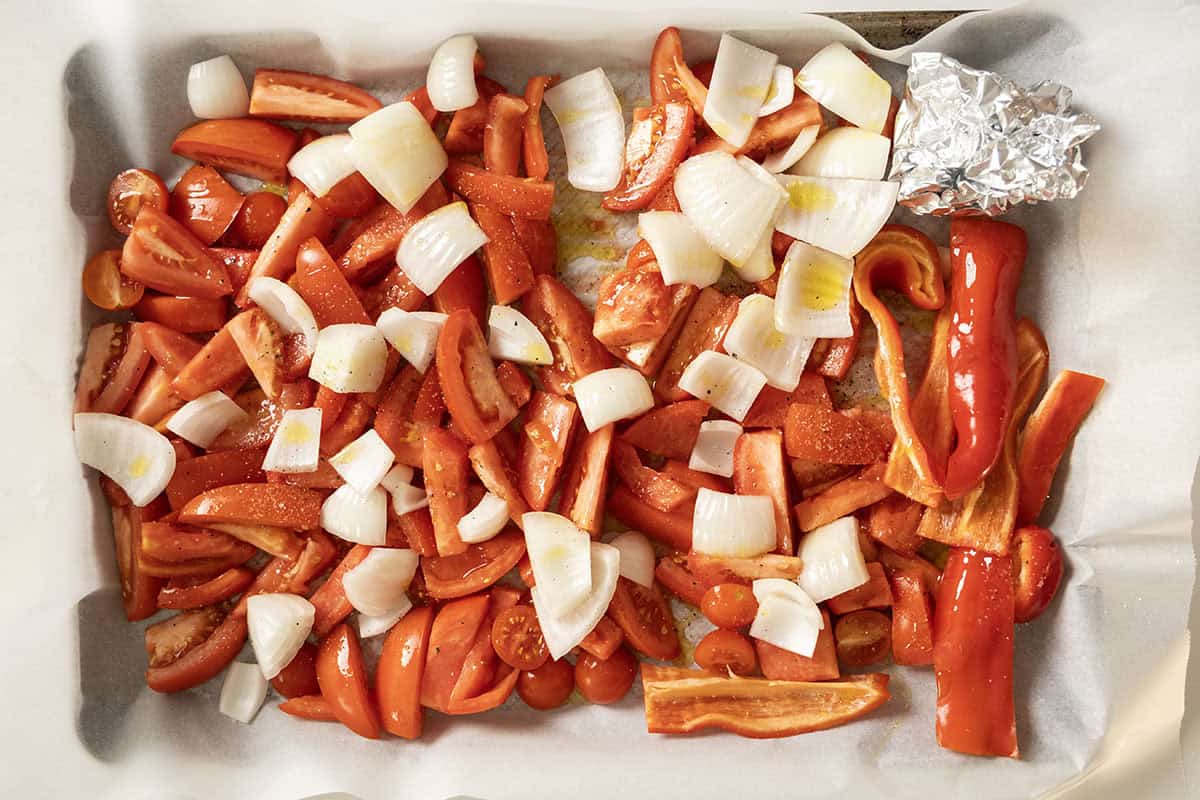 Chopped tomatoes, onions, red pepper, and garlic in aluminum foil on a baking sheet.