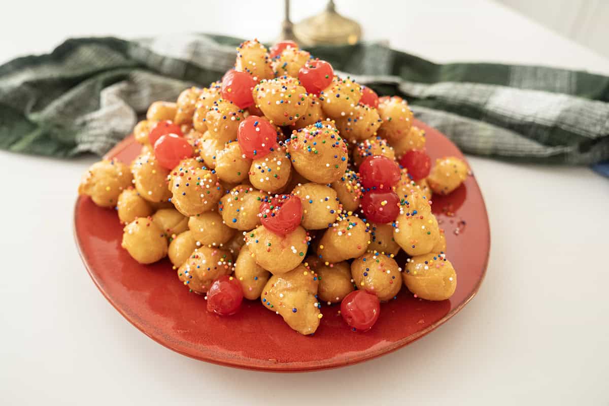 Struffoli piled into a pyramid topped with non-pareils and maraschino cherries.